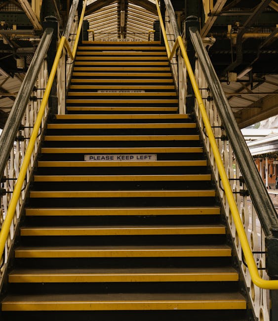 A wide set of metal stairs with yellow safety handrails on each side and black anti-slip strips on each step, leading up to an outdoor platform or upper floor within a warehouse or industrial building. The stairs are enclosed by a metal framework and surrounded by overhead beams and industrial piping. A sign on the middle of the stairs reads 'PLEASE KEEP LEFT,' indicating a movement or safety guideline. The environment has industrial lighting, highlighting the metallic and utilitarian nature of the stairs, which are part of a building used for home relocation or furniture transport logistics. The overall scene emphasizes the structural aspect of stair access within a moving or storage facility, relevant to the context of house removals and managing access challenges such as narrow stairs or lifts, as outlined in the page about access problems for Haringey removals.