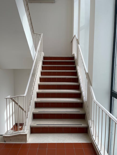 A straight indoor staircase with nine steps, featuring reddish-brown tiled treads and white risers, situated in a well-lit interior space. The staircase is enclosed by white metal railings on both sides, with vertical bars and horizontal handrails. Natural light enters from large windows on the right, illuminating the area. The stairs lead to an upper floor, and a small gap is visible between the bottom of the stairs and the floor, suggesting accessibility considerations. The surrounding walls are painted white, and the landing at the top appears to be spacious. This scene depicts the stairway within a residential building, consistent with house removals and moving services requiring navigation through narrow stairs and lifts, as handled by Removal Companies Haringey.