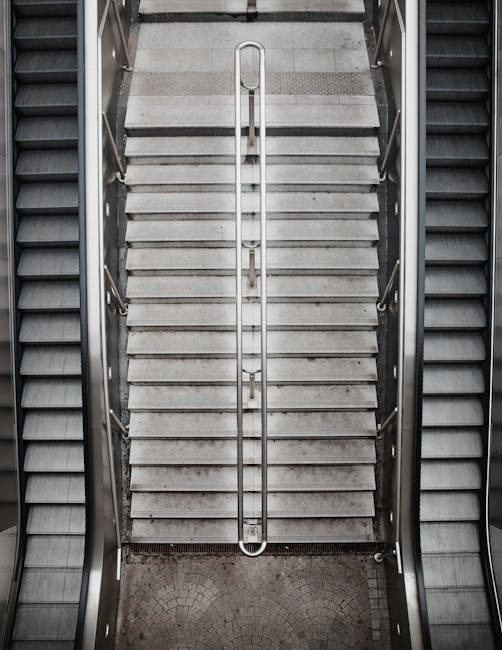 A wide set of metal stairs with yellow safety handrails on each side and black anti-slip strips on each step, leading up to an outdoor platform or upper floor within a warehouse or industrial building. The stairs are enclosed by a metal framework and surrounded by overhead beams and industrial piping. A sign on the middle of the stairs reads 'PLEASE KEEP LEFT,' indicating a movement or safety guideline. The environment has industrial lighting, highlighting the metallic and utilitarian nature of the stairs, which are part of a building used for home relocation or furniture transport logistics. The overall scene emphasizes the structural aspect of stair access within a moving or storage facility, relevant to the context of house removals and managing access challenges such as narrow stairs or lifts, as outlined in the page about access problems for Haringey removals.