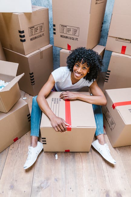 A young woman with curly black hair, wearing a white t-shirt, blue jeans, and white sneakers, is sitting on a wooden floor surrounded by numerous cardboard boxes used for packing household items during a home relocation. She is smiling and holding a large closed cardboard box with red packing tape, which is positioned in front of her. Several other boxes, some open and some sealed, are stacked around her at varying heights, some leaning against a blue wall in the background. The scene depicts a typical house removal process, with the woman preparing items for transportation, fitting into the context of professional furniture transport and moving logistics provided by Removal Companies Haringey. The environment is well-lit, emphasizing the organized nature of packing for a landmark move, as referenced in the Alexandra Palace house removals advice page. The scene highlights the importance of careful packing and handling materials during the packing and moving stage of house removals.