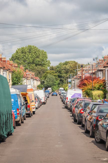 A residential street lined with parked cars on both sides, including compacts, hatchbacks, and vans, with some vehicles covered with protective tarps or blankets. The pavement appears narrow, with a mix of concrete and asphalt, and the street is flanked by traditional terraced houses with brick facades, small front gardens, and varying window styles. Overhead, multiple power lines run parallel to the road, crossing from one side to the other. In the background, there are tall, green trees and a cloudy sky, indicating typical urban residential surroundings. The scene is part of a house removal or home relocation process, where furniture, boxes, or packing materials could be loaded onto a vehicle, possibly operated by Removal Companies Haringey, as part of their removals services. The environment suggests a calm, organized setting for loading furniture and belongings during a house move, with visibility of a loading or parking area extending into a van or moving vehicle at the curb.