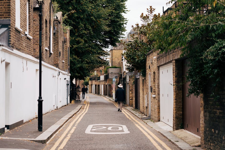 A narrow residential street in Crouch End N8 lined with brick houses and trees, with a speed limit painted as '20' on the asphalt. The street features a double yellow line along the curb and a sidewalk with a black lamp post. In the background, a man dressed in dark clothing and a white hat walks along the pavement, while another person is further down the street. On the right side, adjacent to the houses, there are garage doors, some of which are wooden and others metal, with greenery overhanging from the garden areas. The street scene captures the quiet, urban environment typical for house relocations and furniture transport activities carried out by Removal Companies Haringey, with the arrangement suggesting a space suitable for loading or unloading during home relocation or packing and moving processes, set in daylight with diffuse overcast lighting.
