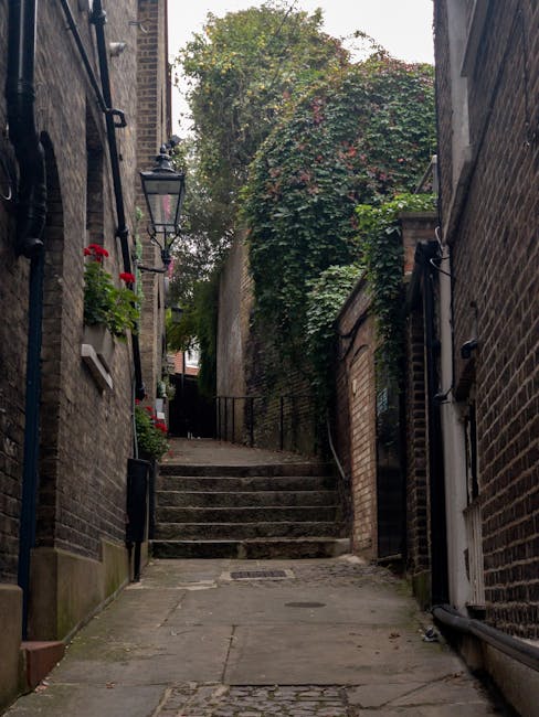 A narrow, ascending street between brick buildings with cobbled paving and stone steps at the top, shaded by trees and surrounded by greenery. On the left side, flower boxes with vibrant red and pink flowers are attached to the buildings, while on the right, a white door with black metal fixtures is visible. The scene depicts a typical urban alleyway that might be encountered during a home relocation or furniture transport process, relevant to services offered by Removal Companies Haringey as part of their house removals in the Crouch End N8 area. The lighting is natural, with daylight filtering through foliage, highlighting the textured brick walls and the ascending staircase, creating a quiet and characteristic street scene suitable for navigating a residential move in a historic or densely built neighbourhood.
