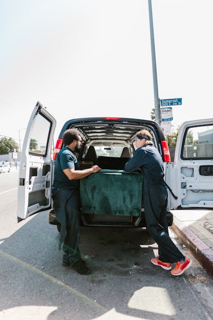 Two professional removalists from Removal Companies Haringey are engaged in a home relocation process, loading a large piece of furniture covered in dark green protective fabric into the rear of a white panel van parked on a pavement near Finsbury Park station. The van's side and rear doors are open, revealing an interior space equipped for furniture transport. One worker, wearing a navy uniform and safety glasses, is holding the furniture steady, while the other, dressed similarly, is assisting with the lifting and positioning. The scene takes place during daylight with sunlight casting shadows on the ground. In the background, street signs and a lamp post are visible. This image illustrates the careful handling and packing of household items as part of a professional removal service, supporting efficient furniture transport and moving logistics typical of house removals in N4, as managed by Removal Companies Haringey.
