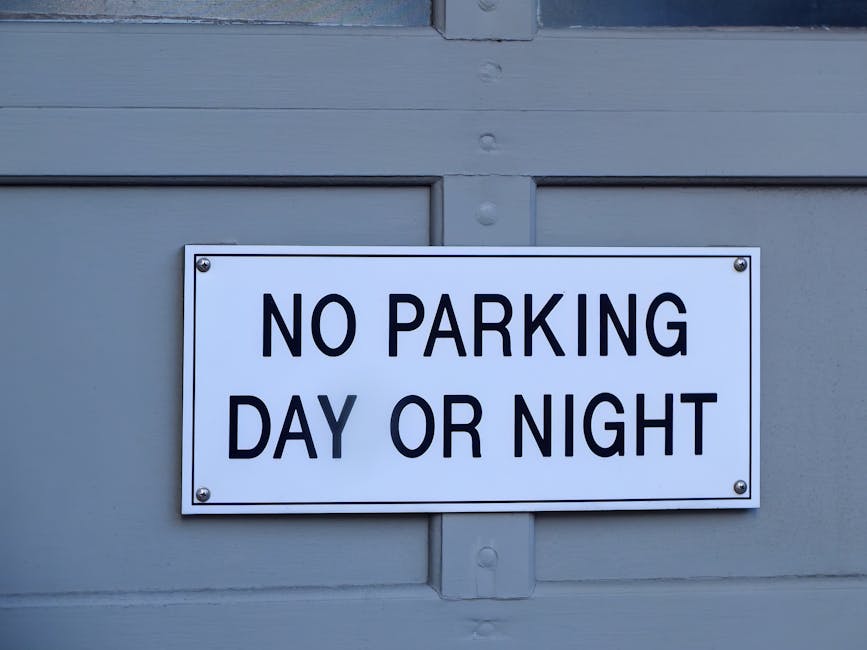 A white rectangular sign with black uppercase text that reads 'NO PARKING DAY OR NIGHT' is mounted on a light gray wooden surface, likely part of a building or garage door. The sign is affixed with four metallic screws, one in each corner. The surrounding area shows smooth painted wood panels and a horizontal wooden beam at the top. This signage indicates parking restrictions potentially relevant to home relocation and furniture transport services with strict adherence to parking rules during moves. The environment appears to be outdoors in a residential or commercial area, with natural daylight illuminating the scene. The chair and boxes, if present in the moving process, are not visible here, but the sign emphasizes the importance of parking compliance during loading and unloading stages. Removal Companies Haringey, operating within the realm of removals and moving logistics, must consider such parking restrictions when planning home relocations in accordance with Haringey Council parking rules.