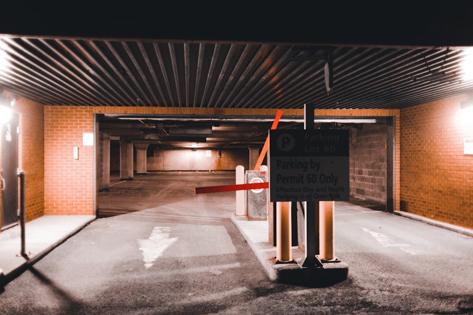An underground parking garage with brick walls and a dark, metal slatted ceiling. The entrance is partially blocked by a parking barrier with red arms in a horizontal position, indicating the parking lot is closed or restricted. On the right side, there is a signpost displaying parking regulation information, including a black parking sign with white lettering and details about permits, relevant to Haringey Council parking rules affecting removals and permits, as noted on the webpage. The paved ground has directional arrows painted in white, guiding vehicles, and the area is illuminated by wall-mounted lights, casting soft light across the concrete floor. Inside the garage, there are multiple concrete support columns, and the space appears ready for furniture transport and home relocation activities, consistent with the services provided by Removal Companies Haringey. The environment reflects a typical urban parking facility suitable for loading and unloading furniture and household items during the moving process.