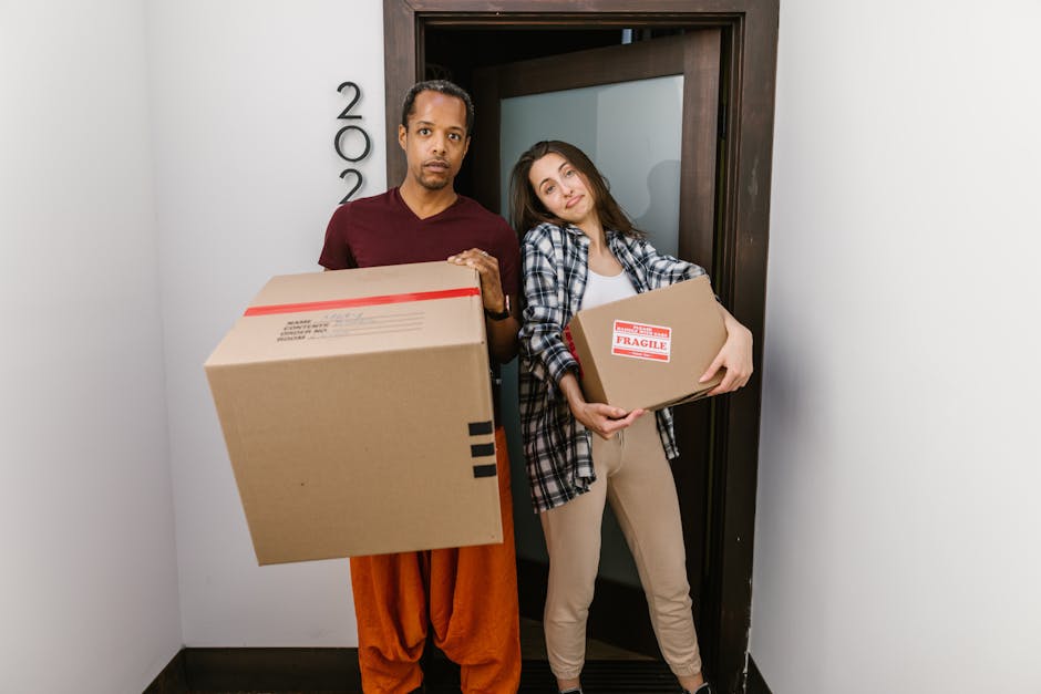 A man and a woman standing inside a residential building at the doorway of a property, each carrying a cardboard box as part of a home relocation process. The man, dressed in a maroon shirt and orange trousers, holds a large box wrapped with red and black tape, while the woman, wearing a plaid shirt and beige trousers, holds a smaller box marked with a red 'FRAGILE' sticker. The doorway behind them features a dark wooden frame, and the wall beside it is white with house numbers visible. The scene is lit with natural or indoor lighting, capturing the unloading or packing process involved in furniture transport and house removals. This image is associated with Removal Companies Haringey, specializing in removals and moving services, and exemplifies the careful handling and logistics involved in packing and loading furniture for a move in Haringey.