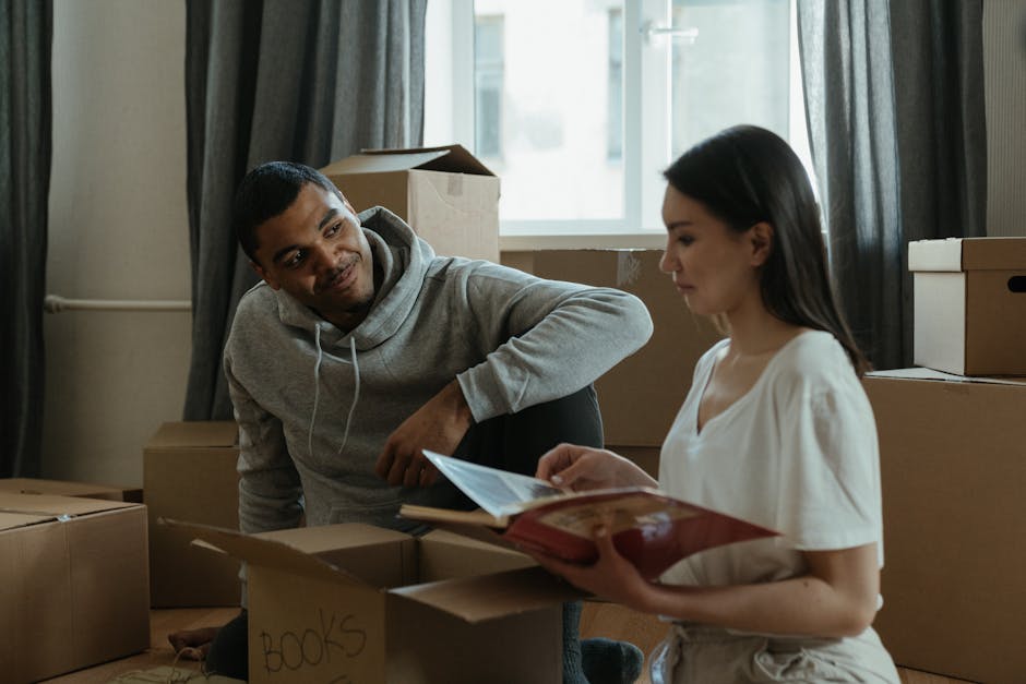 Inside a home during a furniture transport process, a young woman with dark hair wearing a white top is holding an open booklet or packing list while seated among several cardboard moving boxes, some labeled 'BOOKS.' A man with short dark hair, dressed in a grey hoodie, is crouching nearby, looking at the woman and the packing list. The boxes are arranged on the floor near a window with daylight streaming in, illuminating the moving scene. Behind them, additional cardboard boxes and packing materials are visible, indicating preparations for a house relocation undertaken by Removal Companies Haringey. The environment appears tidy, with typical household items and packaging materials, and the action suggests ongoing packing or loading activities as part of the home removal process.