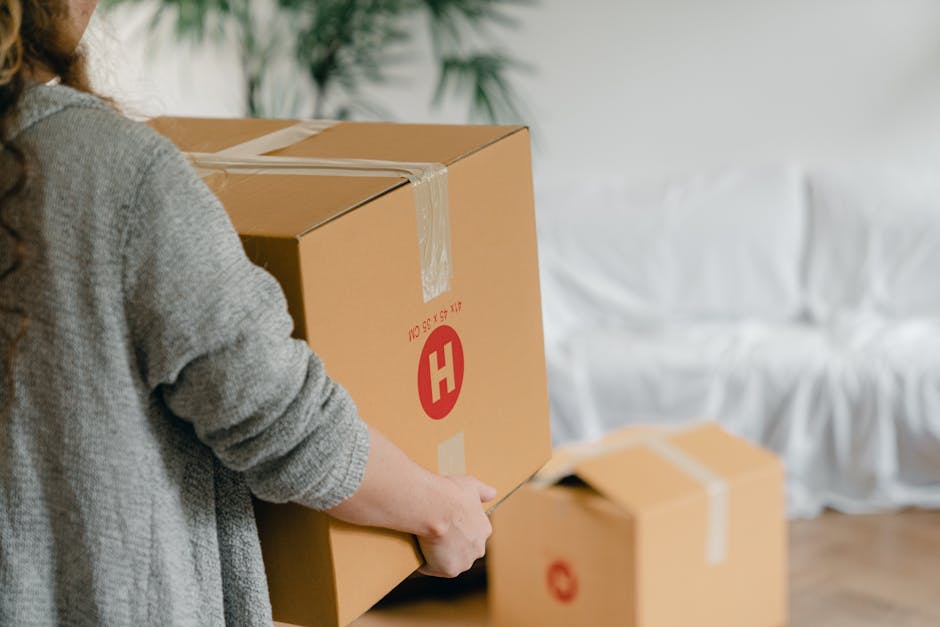 A man and a woman standing inside a residential building at the doorway of a property, each carrying a cardboard box as part of a home relocation process. The man, dressed in a maroon shirt and orange trousers, holds a large box wrapped with red and black tape, while the woman, wearing a plaid shirt and beige trousers, holds a smaller box marked with a red 'FRAGILE' sticker. The doorway behind them features a dark wooden frame, and the wall beside it is white with house numbers visible. The scene is lit with natural or indoor lighting, capturing the unloading or packing process involved in furniture transport and house removals. This image is associated with Removal Companies Haringey, specializing in removals and moving services, and exemplifies the careful handling and logistics involved in packing and loading furniture for a move in Haringey.