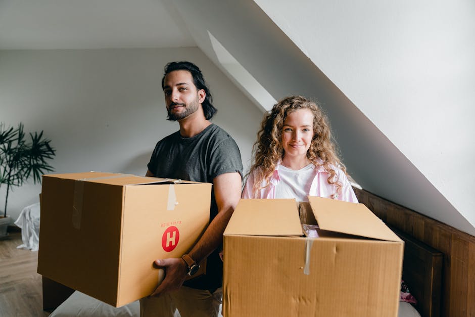 A man and a woman stand inside a room with white walls and a sloped ceiling, each holding a large cardboard box with packing tape. The man has dark hair, a beard, and is wearing a grey t-shirt, while the woman has curly blonde hair and is dressed in a white top with a pink jacket. They are positioned near a wooden headboard and a window, with natural light illuminating the space. In the background, there is a potted plant and some partially visible furniture, indicating an active home relocation or packing and moving process. The boxes are plain brown cardboard, some wrapped with packing tape, and are of varying sizes, suggesting the process of loading and transporting belongings. The scene captures a household in the midst of a furniture transport or packing phase, exemplifying the services offered by [COMPANY_NAME] for efficient removals in Haringey.