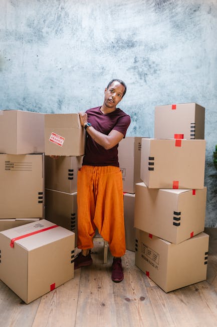 A man standing in a room surrounded by numerous cardboard boxes of various sizes, some stacked on top of each other and others placed on the wooden floor. The boxes are sealed with red and black tape, indicating they are packed for a home relocation. The man is wearing a maroon T-shirt and orange trousers, and is leaning against one of the boxes with one arm, looking directly at the camera. The room has a light blue textured wall and natural lighting, suggesting a setting where packing and moving preparations are underway, consistent with professional removals services provided by Removal Companies Haringey. The scene captures the process of furniture transport and packing during a house move, highlighting the company's role in managing logistics for home relocations.