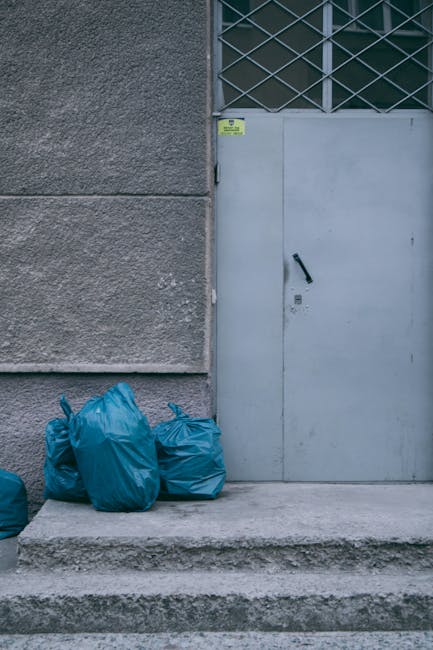 Several blue garbage bags filled with household waste are placed on the concrete steps outside a building near a metal door with a handle and locking mechanism. The building wall is textured and grey,