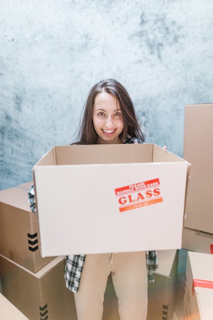 A woman with long brown hair and a cheerful expression is standing indoors in front of a light blue textured wall, holding an empty cardboard moving box labeled 'Glass' with red and white tape around the top. She is wearing a plaid shirt over a light-colored outfit. Surrounding her are several other cardboard boxes, some sealed with packing tape, stacked on the floor and against the wall. The scene depicts a packing and moving process associated with house removals, with the woman preparing for a home relocation. The area is well-lit, and the boxes are in various sizes, indicating an organized packing effort in the context of furniture transport and moving logistics, as carried out by companies like Removal Companies Haringey.